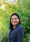A young asian woman with short dark brown hair wearing a dark gray suit and glasses smiles at the camera in front of a lush green background.